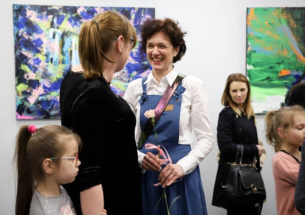 Na zdjęciu widzimy uśmiechniętą Monikę Marek na otwarciu swojej wystawy w Galerii Zajezdni Kultury w Pleszewie. Monika trzyma goździk w kolorze białym. Obok stoi kobieta z dzieckiem. W tle obrazy artystki.