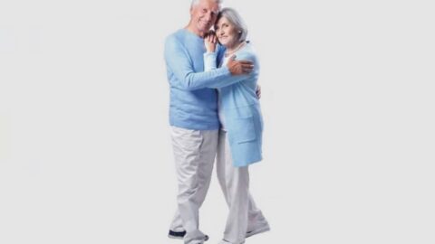 Grafika przedstawia na szarym tle stojące dwie osoby, kobietę i mężczyzneę w objęciu, uśmiechnięci, patrzący do w naszym kierunku. Oboje są seniorami z siwymi włosami. Ubrani są w jednolity strój: białe spodnie i błękitna góra w postaci koszulki u mężczyzny i sweterka u kobiety.