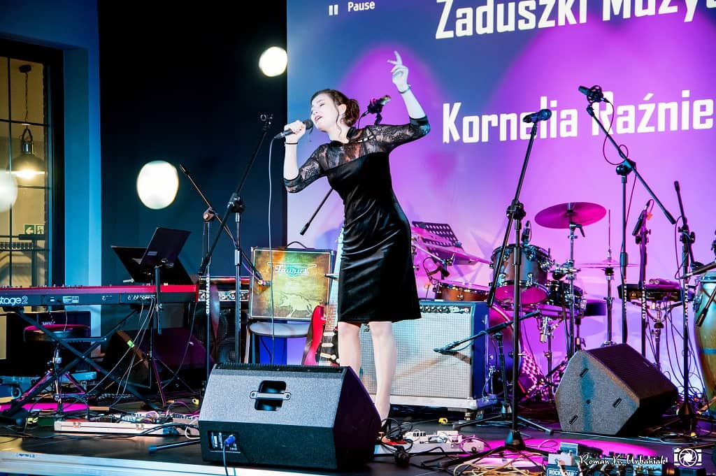 Zdjecie przedstawia występ Kornelii Raźniewskiej ubranej w czarną stylową sukienkę w ekpresyjnej pozie wokalnej podczas koncertu Zaduszki Muzyczne odbywające się w Zajezdni Kultury w Pleszewie