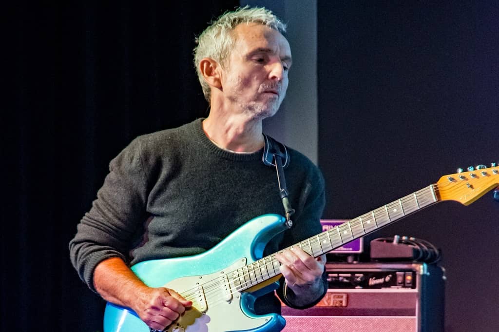 Zdjecie przedstawia koncert Apostolisa Anthimosa ubranego w czarną koszulkę i grającego na błękitnej gitarze Fender Stratocaster w Zajezdni Kultury w Pleszewie